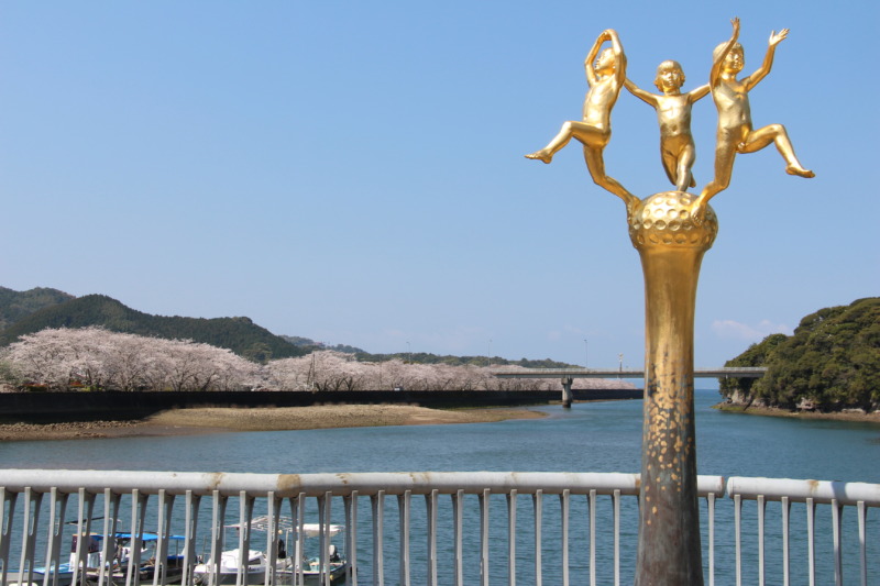 空と山と川を背景に撮影された津奈木町内の彫刻作品（「風ん子」岩野勇三）
